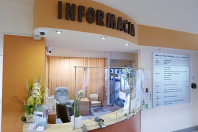 Zdjęcie frontu szpitalnej recepcji. Z jej lewej strony umieszczona jest tablica z informacjami o tym, co znajduje się na poszczególnych poziomach szpitala.