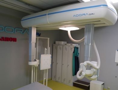 Zdjęcie przedstawia fragment urządzenia do rentgenodiagnostyki, w tle widoczny fartuch ochronny dla wykonującego badanie. 