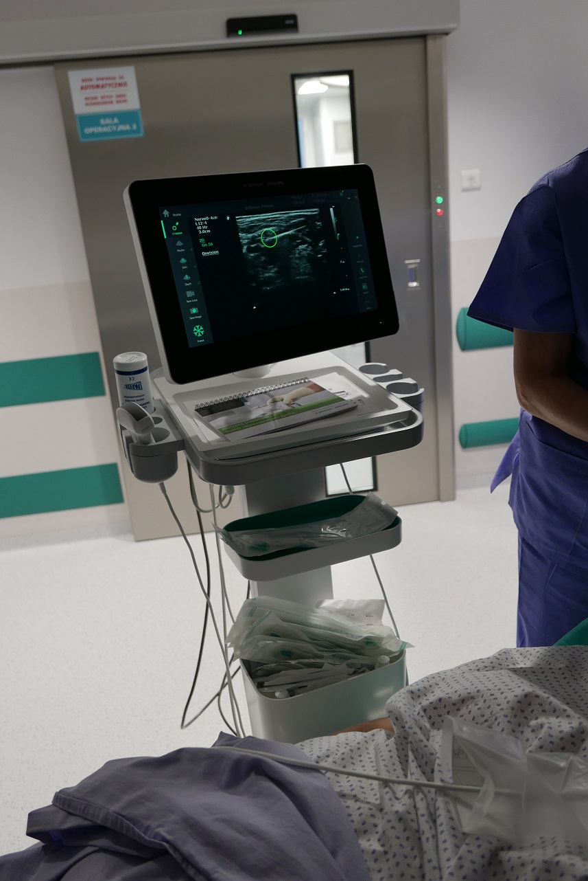 Zdjęcie przedstawia ultrasonograf pracujący podczas procedury znieczulania. Widoczny jest ekran dotykowy z wyświetlonymi danymi, pionowy korpus urządzenia oraz kable umożliwiające nawigację igłami. 